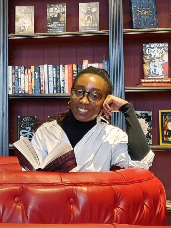 Jeune femme noire portant des lunettes tenant un livre ouvert et posant devant une bibliothèque